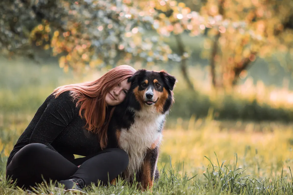 Besitzerin eines black tri Australian Shepherds sitzt mit ihrem Hund auf einer Wiese und lehnt sich an ihn an