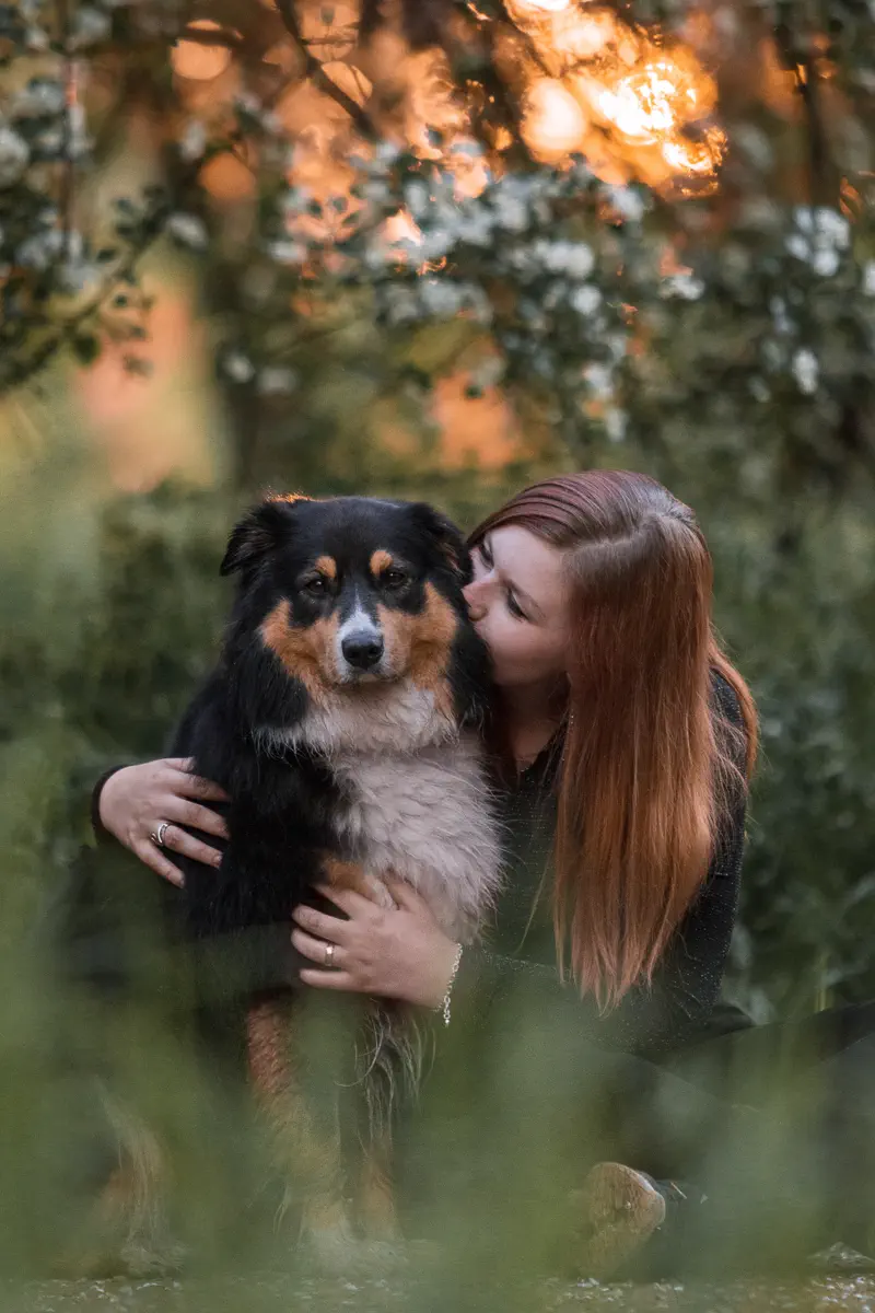 Besitzerin eines black tri Australian Shepherds sitzt neben ihrem Hund, umarmt ihn und gibt ihm ein Küsschen